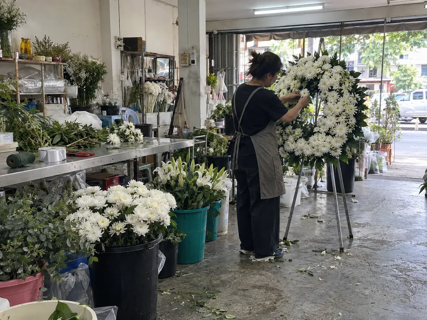 ช่างจัดดอกเบญจมาศสีขาวลงบนโครงพวงหรีดในร้านดอกไม้ มีถังดอกไม้สดวางข้างโต๊ะ ถ่ายจากด้านข้าง