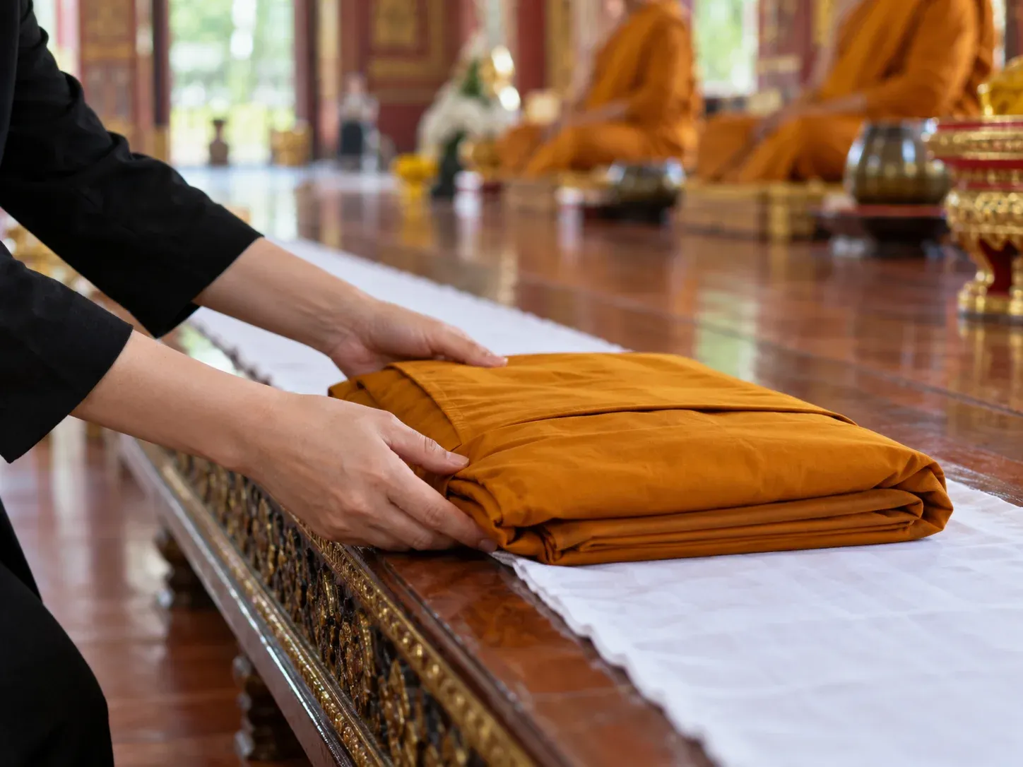 มือผู้หญิงชุดดำประคองผ้าไตรสีเหลืองด้วยสองมือ กำลังวางลงบนแถบผ้าขาว