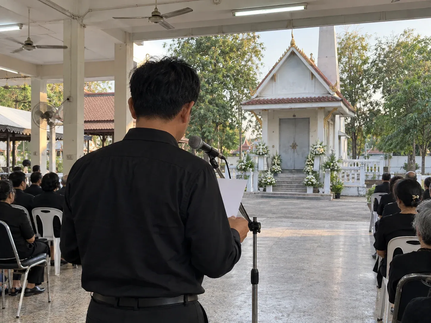 ชายชุดดำยืนหน้าไมโครโฟนถือกระดาษ มองจากด้านหลัง หน้าเมรุวัดไทยที่ประดับดอกไม้ขาว