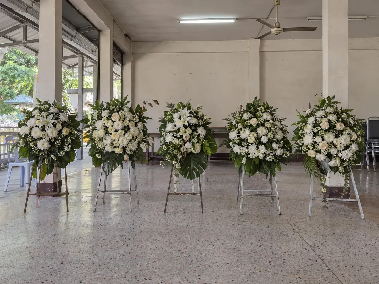 พวงหรีดดอกไม้สดสีขาวหลายพวงตั้งเรียงบนขาตั้งริมผนังศาลาวัด