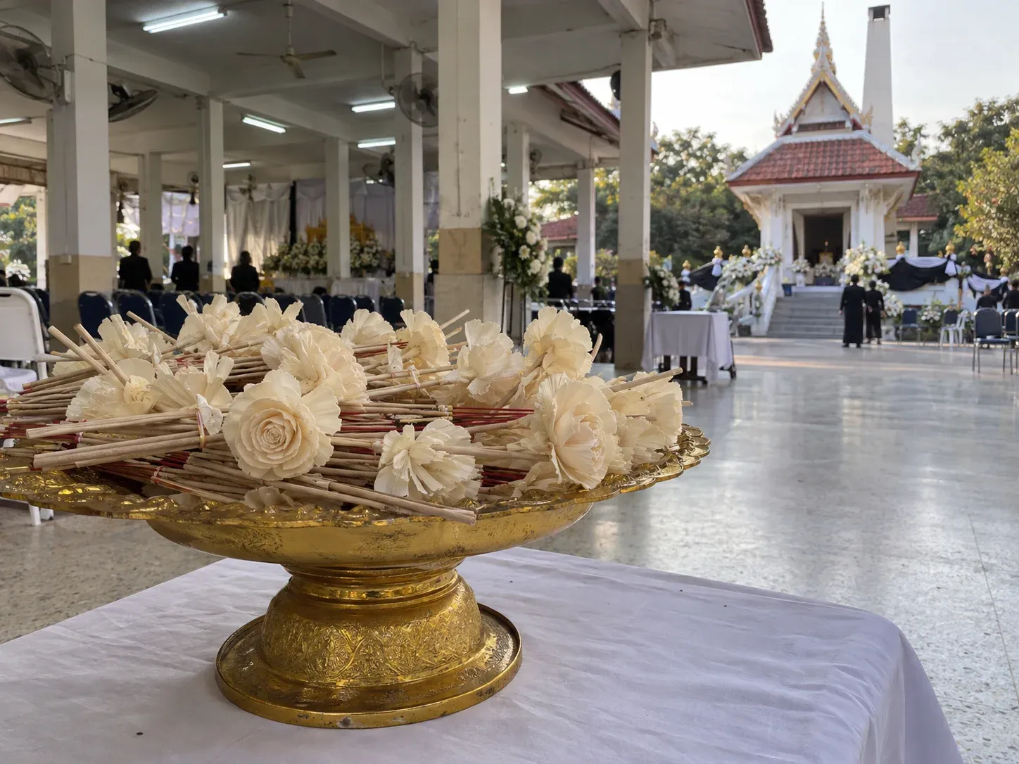 พานดอกไม้จันทน์สีครีมวางบนโต๊ะผ้าขาว หน้าเมรุวัดไทยช่วงเย็น