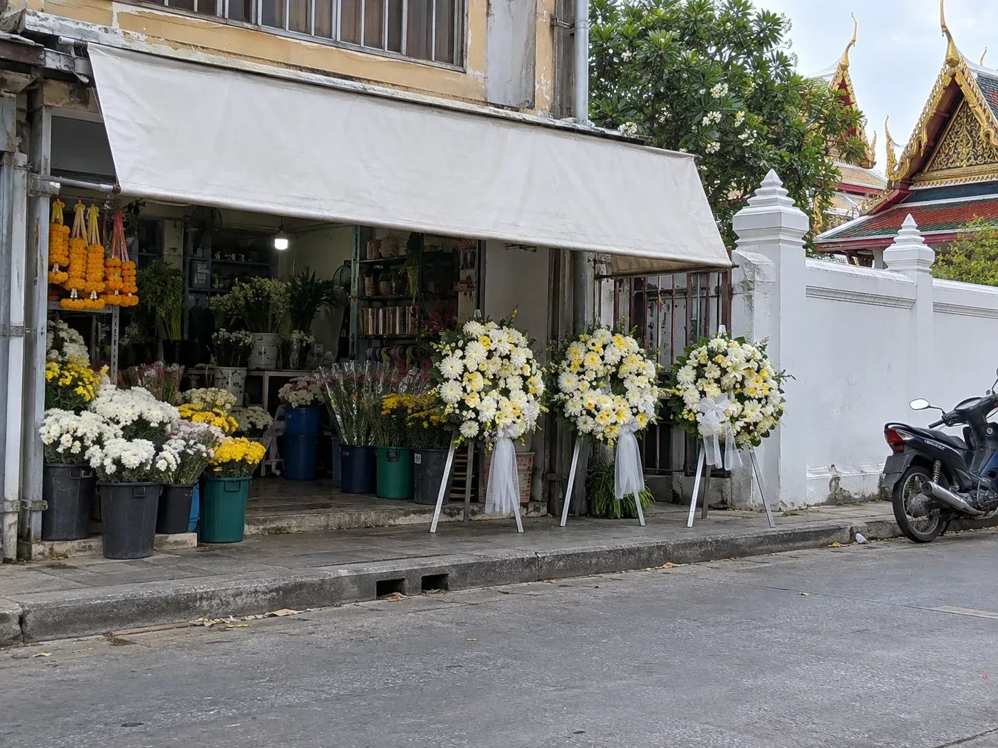 ร้านดอกไม้เล็กๆ ริมถนนใกล้กำแพงวัด มีพวงหรีดดอกไม้สดตั้งโชว์หน้าร้านและถังดอกไม้สด