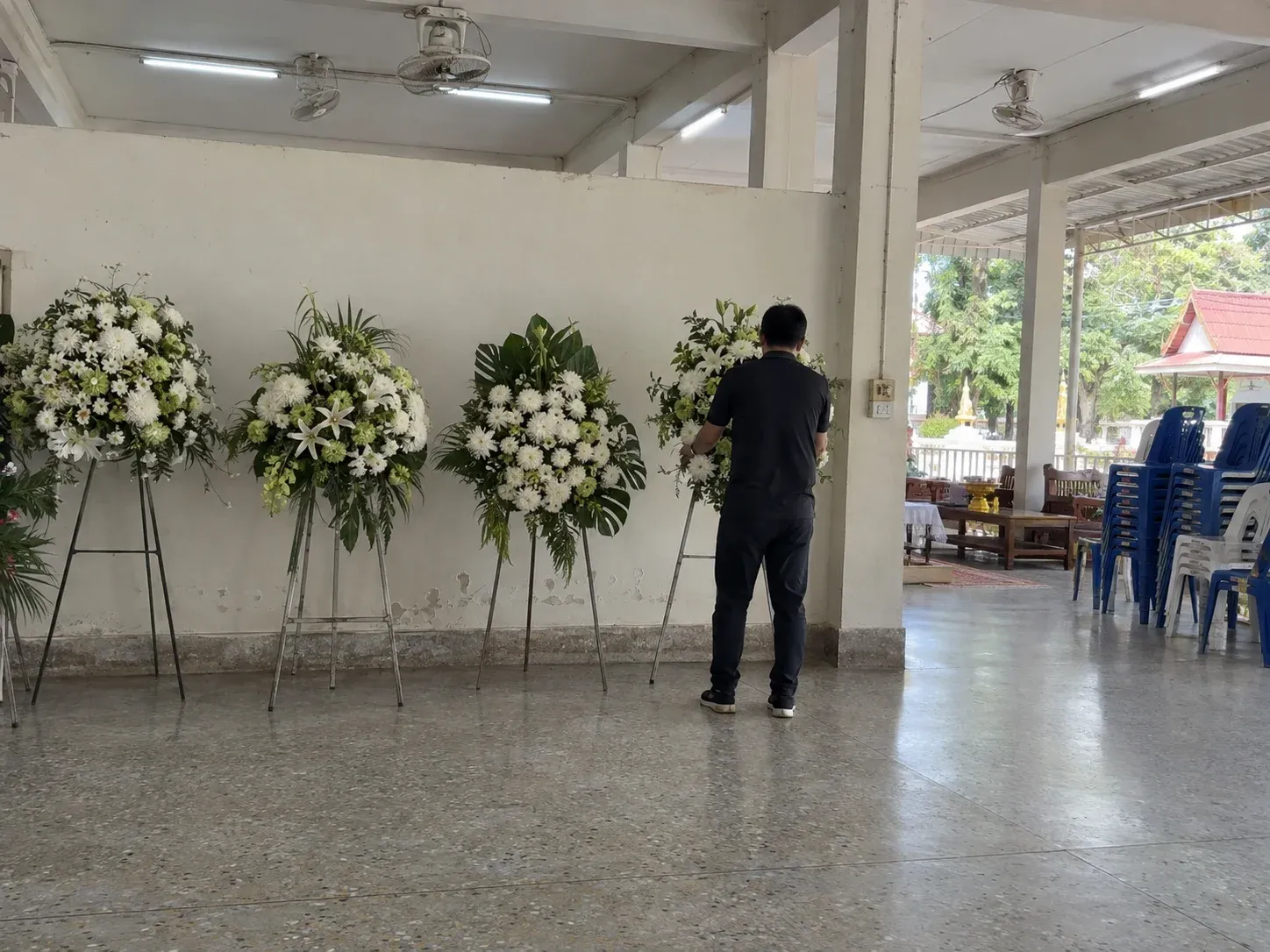 พนักงานตั้งพวงหรีดดอกไม้สดบนขาตั้งเรียงต่อจากพวงหรีดอื่นหน้าศาลาวัด ถ่ายจากด้านหลัง